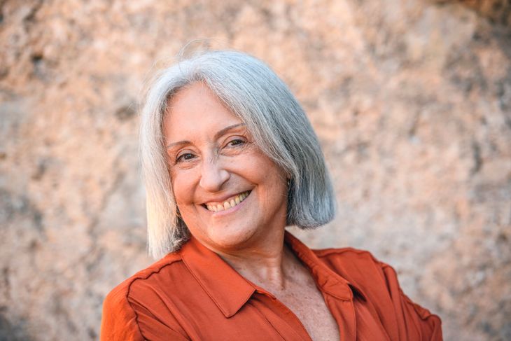 Smiling 69 year old woman with gray bob wearing rust colored open collar blouse and smiling at camera with arms crossed.