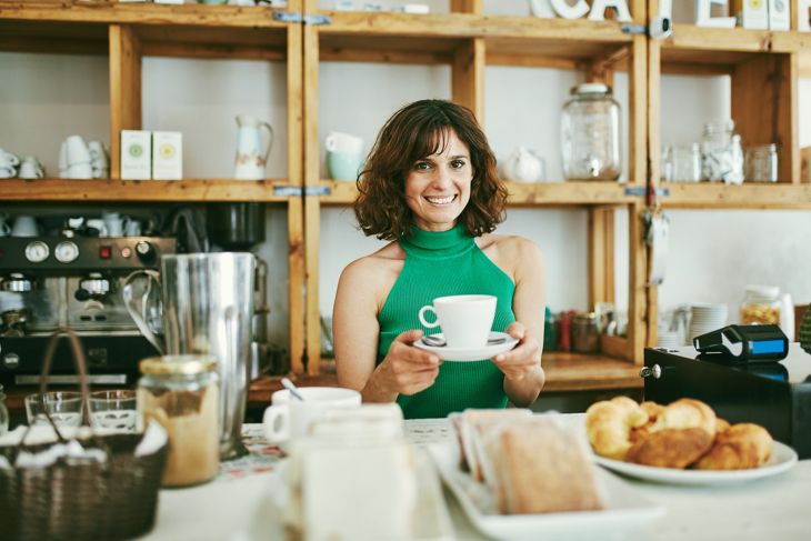 Front view of Hispanic female worker in early 40s smiling at camera as she stands ready to serve cup of coffee behind counter in Buenos Aires cafe.