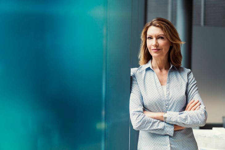 Portrait of confident businesswoman standing with arms crossed. Female professional is wearing smart casuals. She is leaning on blue wall at office.