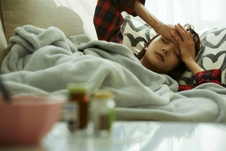 Close-up of a sick woman resting on heart-patterned pillows with her eyes closed. A person in a red plaid shirt places a hand on her forehead to check her temperature. Medicine bottles and a bowl sit on a table in the foreground.