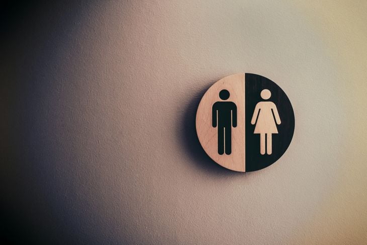 A minimalist, circular restroom sign mounted on a textured wall. The sign is divided down the middle with a light wood left side and a black right side, featuring contrasting male and female symbols.