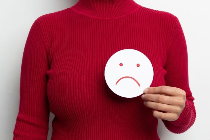 A person in a red turtleneck holds a round paper with a sad face over their chest, symbolizing the common experience of breast cysts, which can range from harmless to uncomfortable and may require monitoring or further tests. 