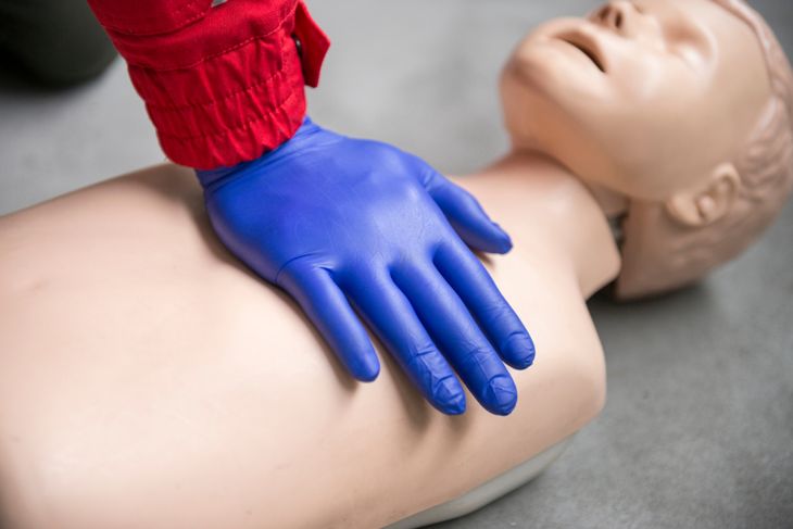 A close-up of a gloved hand in a blue nitrile glove performing chest compressions on a child-sized CPR manikin, showing proper hand placement for pediatric CPR. 