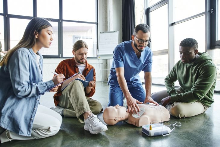 A CPR instructor kneels beside a training manikin, demonstrating pad placement for an AED while three adults watch and take notes in a well-lit classroom, emphasizing how to respond safely and quickly in emergencies. 