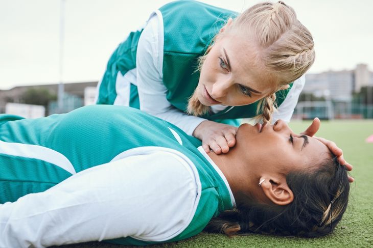 A soccer player in a green uniform tilts back a teammate’s head on the field, preparing to give rescue breaths during an emergency. 