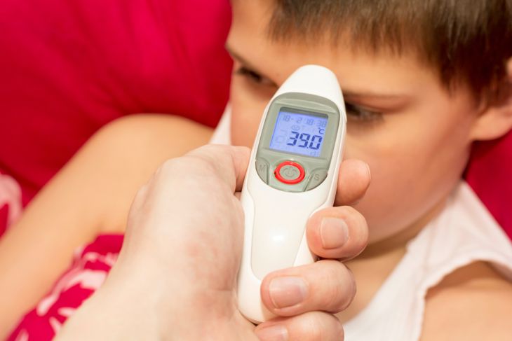 A close-up of a child in bed as an adult holds a digital thermometer showing a high temperature of 39°C, highlighting that sudden high fever is an early sign of Naegleria fowleri infection. 