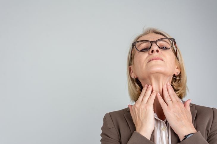 An older woman wearing glasses tilts her chin up and gently feels both sides of her neck, demonstrating how swollen lymph nodes can be checked for changes—an important step in monitoring for infection or other health concerns. 