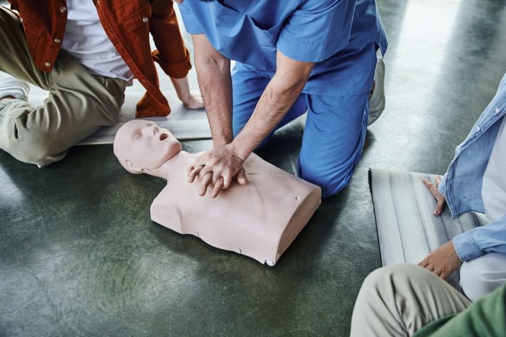 A close-up of a person in blue scrubs performing chest compressions on a CPR manikin, with others observing, demonstrating the proper technique and importance of effective compressions during CPR. 