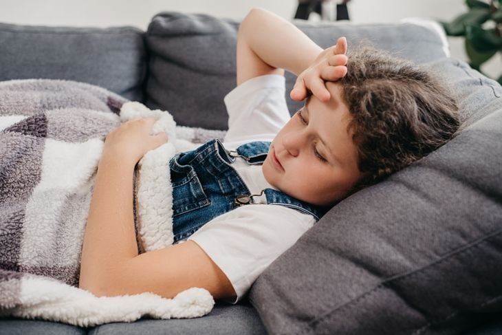 A young girl lies on a couch under a blanket, holding her forehead and appearing uncomfortable, suggesting a severe headache that can be an early symptom of Naegleria fowleri infection. 