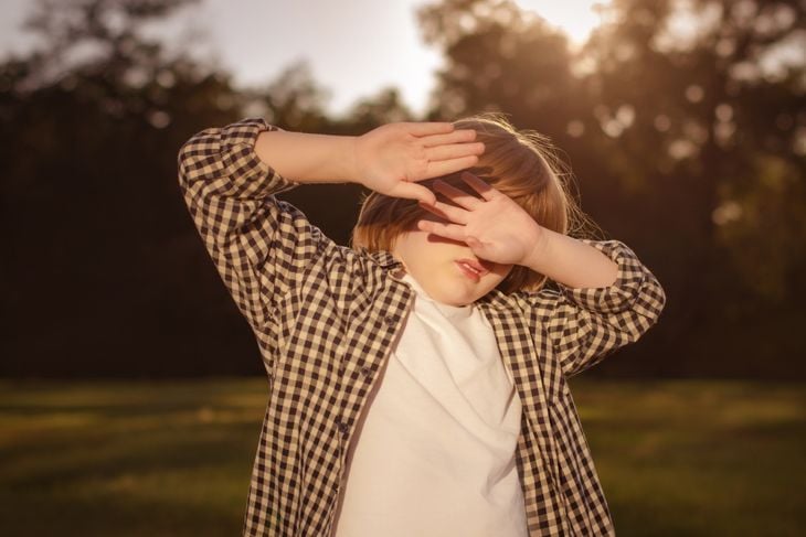 A young child outdoors raises both hands to shield their eyes from bright sunlight, suggesting light sensitivity, which, along with neck stiffness, can be an early sign of Naegleria fowleri infection. 