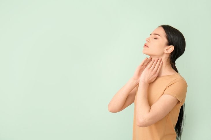 A woman stands against a pale green background with her hands gently touching her neck, demonstrating how nodules can feel like firm lumps in the body, including the thyroid or under the skin. 