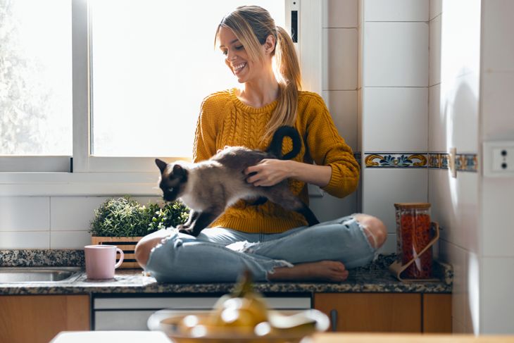 Shot of beautiful youg woman sitting on kitchen table while stroking a cat sitting on her lap at home.