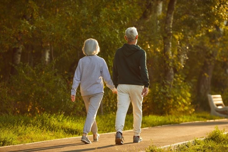 Older adults walking together on a paved path through a park, shown from behind in sneakers and casual clothes, suggesting steady, purposeful movement as part of a regular outdoor routine
