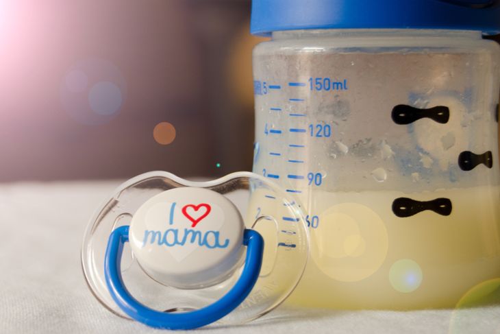 A baby bottle filled with yellowish colostrum sits next to a blue and white pacifier that reads “I ❤️ mama,” resting on a soft surface with a warm lens flare effect in the background.