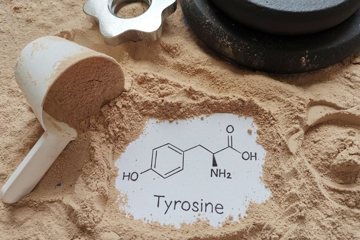 A scoop of protein powder rests on a white surface scattered with powder, partially revealing a card showing the chemical structure and name of tyrosine. Metal gym equipment and weight plates sit nearby, suggesting a fitness or supplement context.