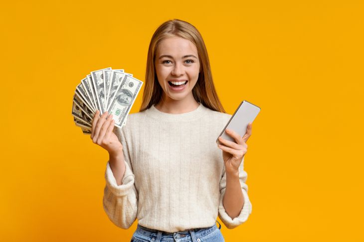 Happy teen girl showing holding smartphone and bunch of money, orange background