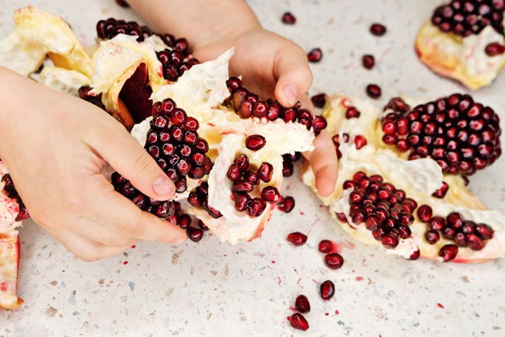 Two hands separate sections of a pomegranate, exposing clusters of deep red seeds still attached to the white pith. Loose seeds are scattered on a speckled countertop.