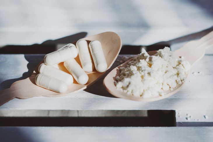 Two wooden spoons rest on a white wooden surface — one holds white capsules, the other contains a pale, powdery substance resembling dried colostrum. Bright natural light casts soft shadows across the scene.