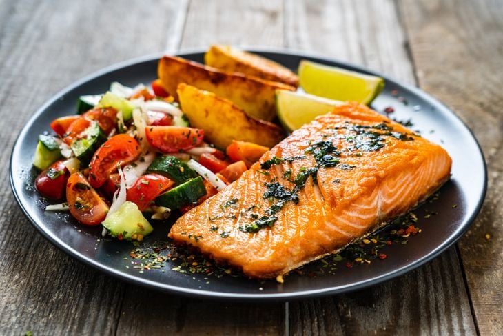 Fried salmon fillet with fried potatoes, lime and vegetable salad served on black plate on wooden table