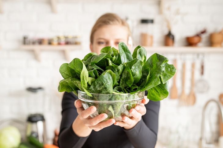Young blond smiling woman holding a bowl of fresh spinach in the kitchen