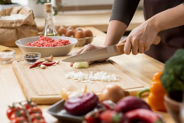 A person chops garlic and onion on a wooden cutting board in a kitchen surrounded by fresh ingredients, including chili peppers, ground meat, eggs, tomatoes, and bell peppers. The focus is on hands and knife technique.