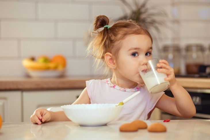 A young girl with her hair in pigtails sits at a kitchen counter drinking a glass of milk. In front of her is a white bowl with a spoon and a few cookies on the counter, with a fruit bowl and jars in the background.