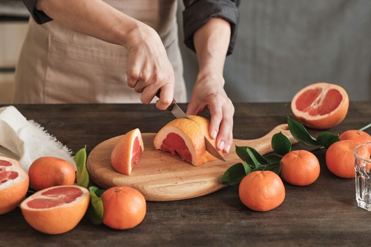 A person in an apron slices a grapefruit on a wooden cutting board, surrounded by whole tangerines with leaves and halved grapefruits. A clear drinking glass and cloth napkin sit nearby on the dark wooden table.
