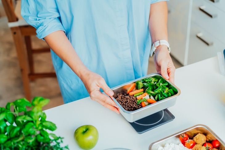 A person in a light blue shirt places a metal meal prep container on a digital scale. The container holds a portion of lentils, sliced carrots, and leafy greens. Nearby are fresh herbs, a green apple, and another prepared dish.