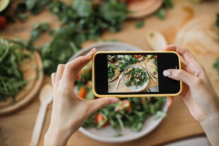 A person holds a smartphone to photograph a fresh arugula salad topped with vegetables. The phone screen displays the same salad plate on a wooden table, surrounded by scattered greens, cutting boards, and wooden utensils.