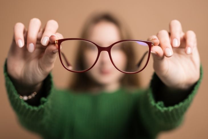 Young girl with poor vision holds glasses with hands forward on beige background