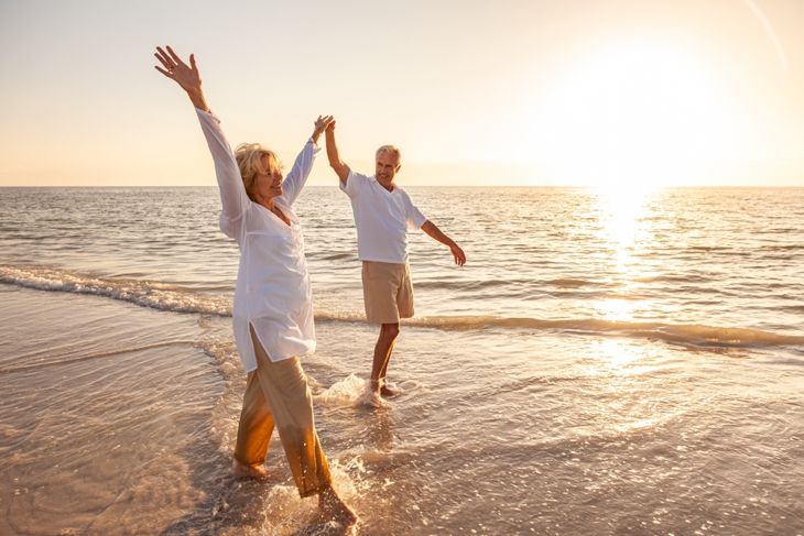 Happy senior man and woman old retired couple walking and holding hands arms raised on a beach at sunset