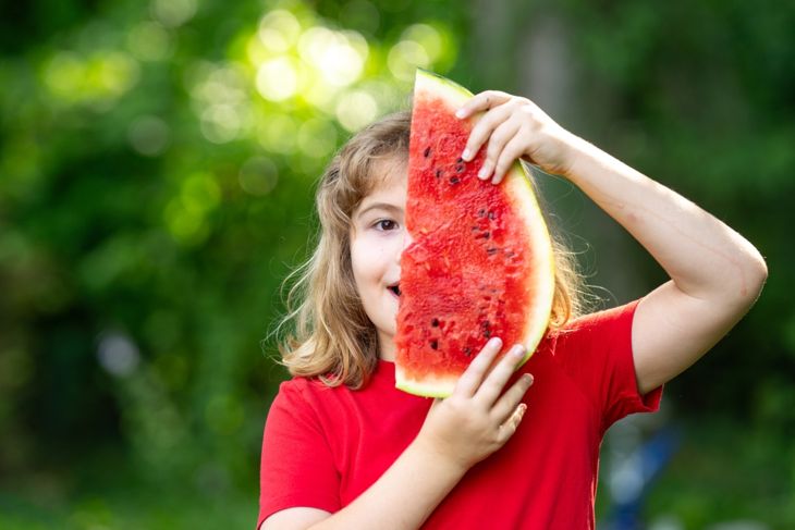 A child in a red shirt holds a large slice of watermelon vertically in front of their face, partially covering one eye. The background is lush and green, suggesting a sunny outdoor setting.