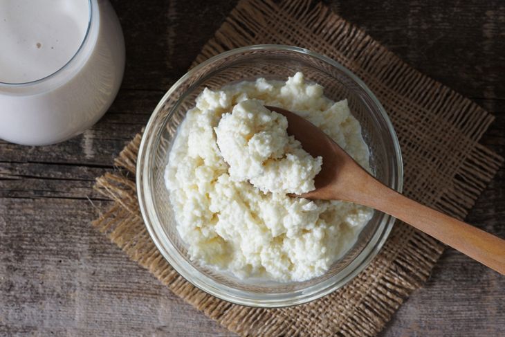 Thank you — here’s revised alt text to reflect that: A glass bowl of raw cow colostrum with a wooden spoon rests on a rustic wooden surface, alongside a glass of milk. The thick, curdled texture and pale color suggest it’s unprocessed and freshly collected.