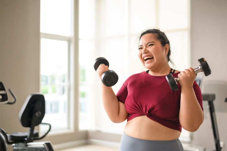 A woman in a red crop top smiles while lifting dumbbells at the gym, with large windows and exercise equipment in the background. Her pose suggests mid-workout movement in a bright, airy fitness space.