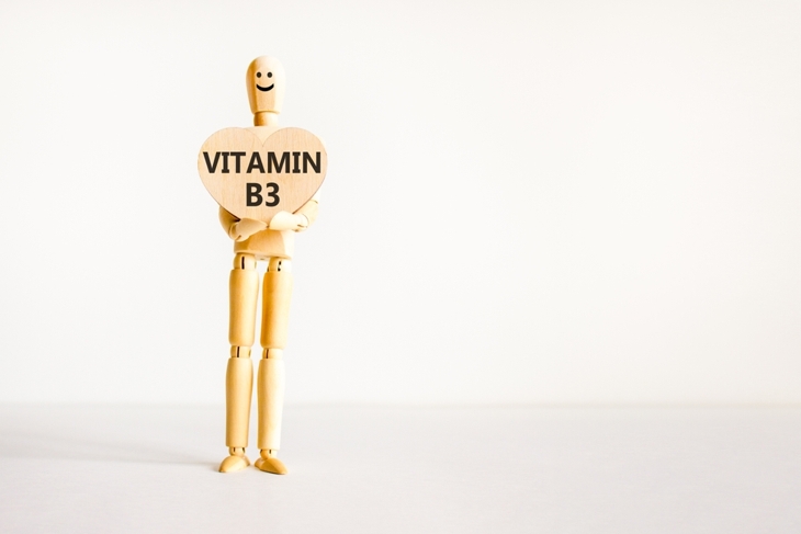 A wooden mannequin with a smiley face holds a heart-shaped sign that reads “VITAMIN B3” against a plain white background. The simple composition emphasizes the message about the vitamin.