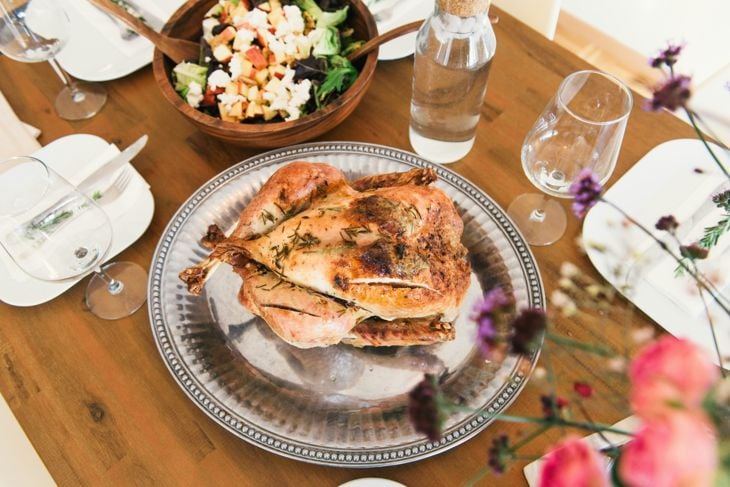 A roasted whole chicken garnished with herbs sits on a silver platter at the center of a wooden dining table. Surrounding it are glasses, white plates with utensils, a wooden bowl of salad with diced fruit and cheese, and a glass bottle of water.