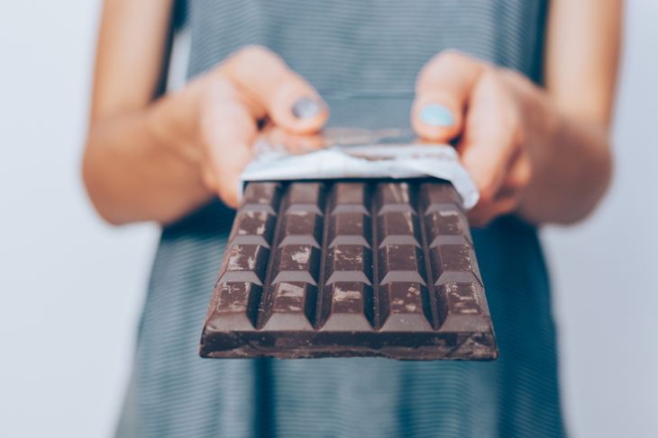 A person in a striped shirt holds out an unwrapped bar of dark chocolate with both hands, offering it toward the camera. The focus is on the chocolate, with fingers and polished nails slightly blurred in the background.