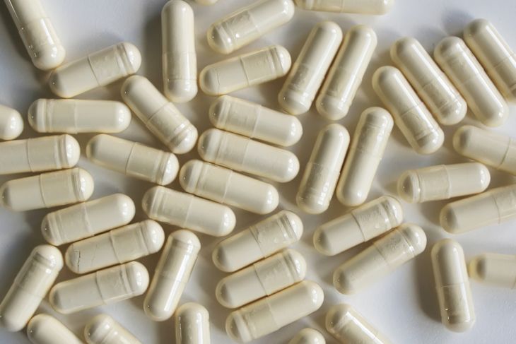 A scattered pile of off-white capsules lies on a smooth white surface. The capsules are uniform in size and shape, with a glossy coating that reflects the light.