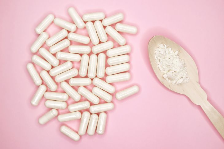 A cluster of white capsules lies on a pink background next to a wooden spoon filled with pale, crumbly powder, suggesting the contents of the supplement in capsule and loose form.