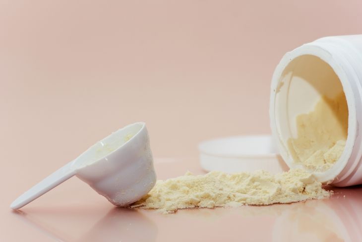 A white plastic container of powdered colostrum lies on its side, spilling pale yellow powder onto a glossy pink surface. A measuring scoop rests nearby, partially filled with the same powder.