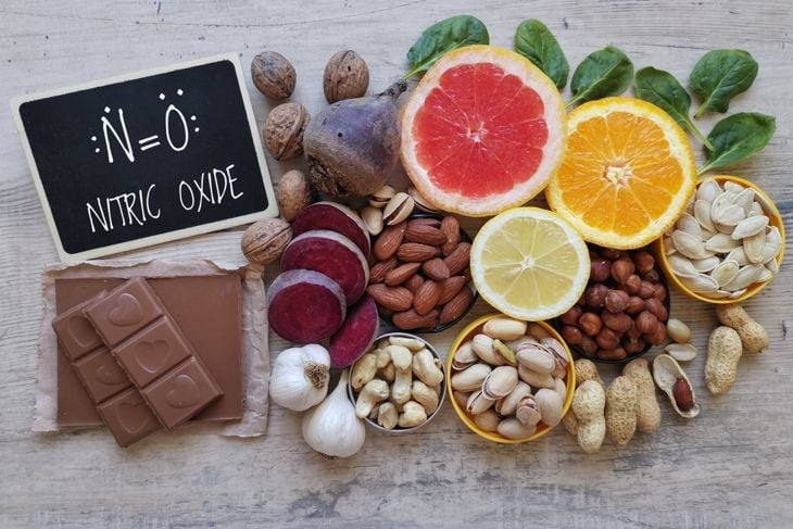 An assortment of nitric oxide–supporting foods is arranged around a small chalkboard with a chemical formula and the words “NITRIC OXIDE.” Items include citrus slices, raw beets, dark chocolate, garlic, nuts, seeds, spinach leaves, and whole walnuts on a wooden surface.