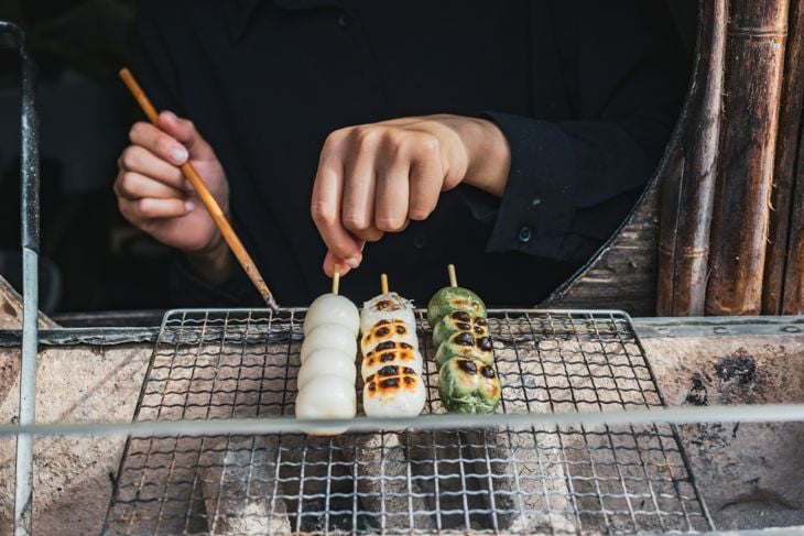 Three skewers with dango being grilled over charcoal on a wire mesh grill held by a person.