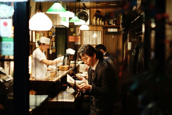 People sitting at a restaurant counter looking at menus, with chefs preparing food in the background.
