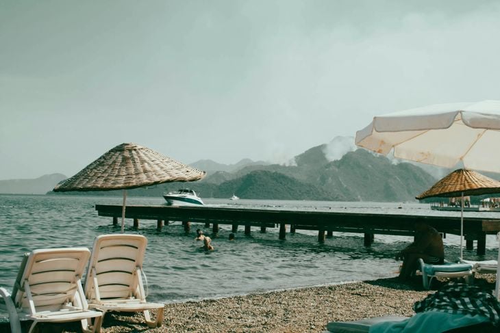 Beach chairs and umbrellas near a calm sea with a wooden pier extending over the water and mountainous background.