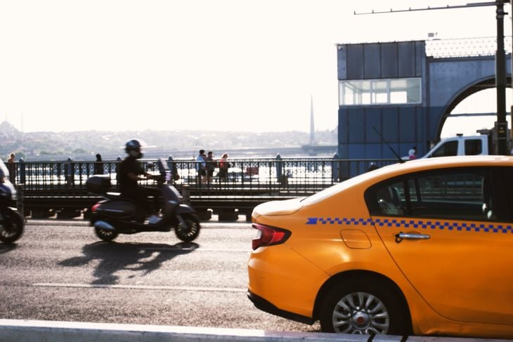 Yellow taxi and motorbikes on a city road with a bridge and buildings in the background.