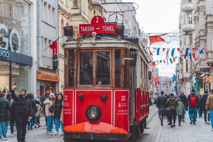 Red vintage tram moving through a crowded street lined with shops and pedestrians under festive flags.