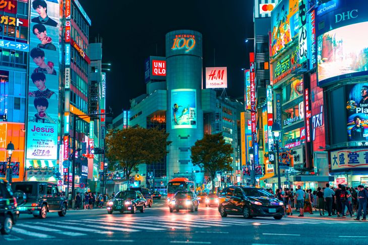 Busy urban street at night with colorful illuminated billboards, cars, and pedestrians crossing the road.