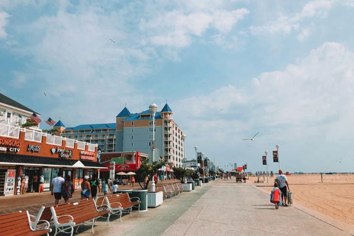 Beach promenade with benches, buildings, and people walking under a partly cloudy sky on a sunny day.