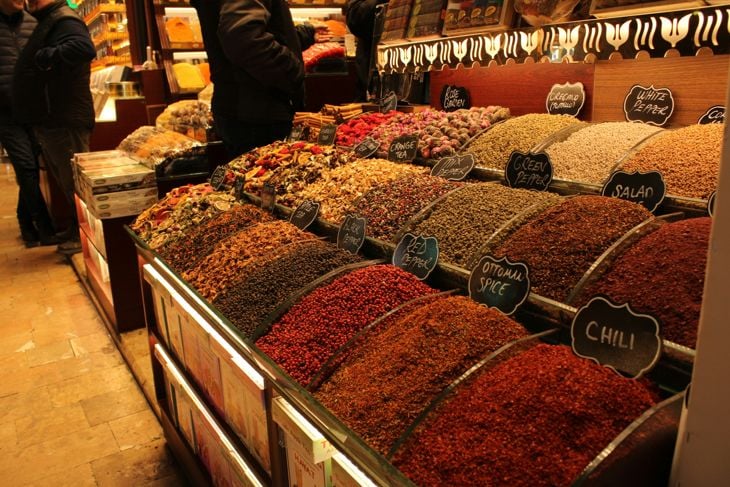Colorful spices and herbs displayed in open containers at a market stall with shoppers in the background.