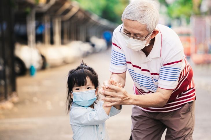 Elderly man and young girl wearing masks, holding hands outdoors on a sunny day.